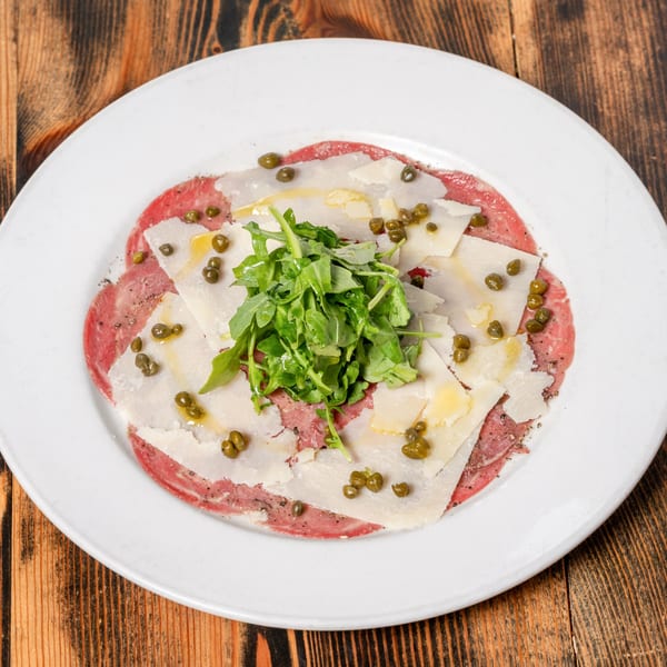 Carpaccio with thinly sliced meat, parmesan, capers, and arugula on a white plate.