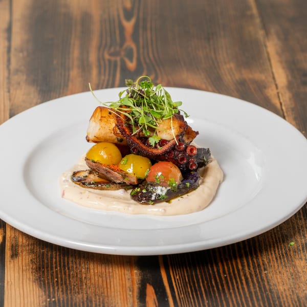 Grilled octopus with cherry tomatoes, mushrooms, and microgreens on a white plate.